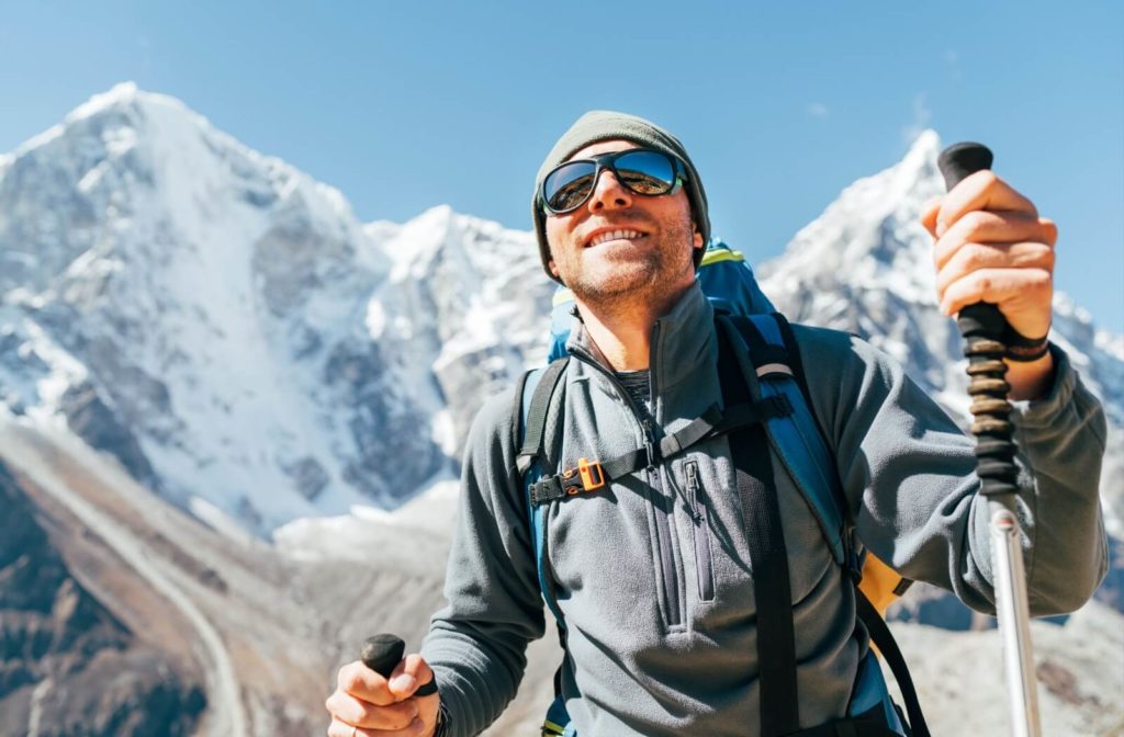 A man on top of a mountain wearing sporty sunglasses and a backpack while hiking with poles.