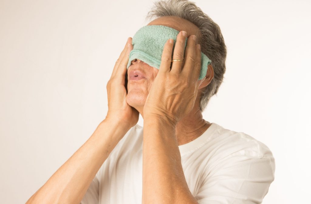 An older man holding a cold compress on his eyes as a home remedy for eye allergies.