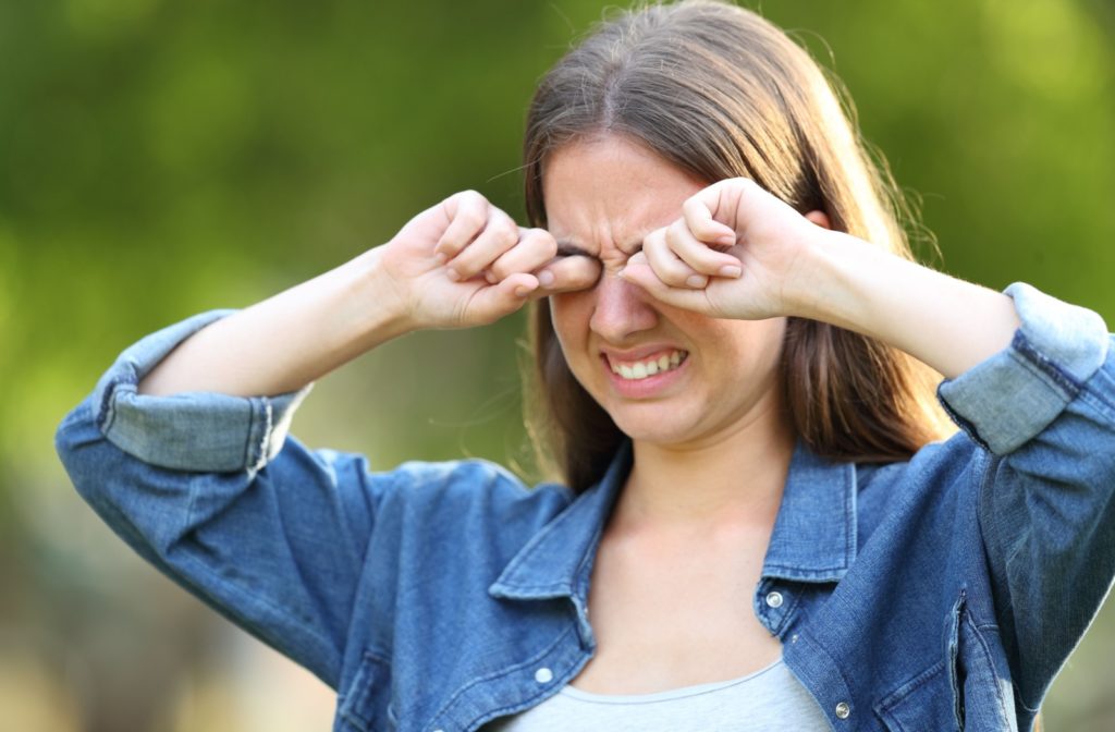 A young woman looks like she is uncomfortable as she rubs her eyes while standing outside.