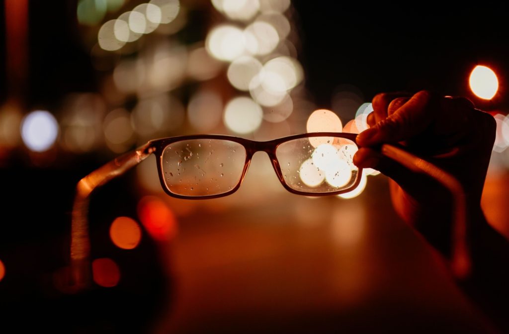 A hand holds up a pair of glasses with a few in-focus lights. Beyond the glasses are blurry lights seen with astigmatism.