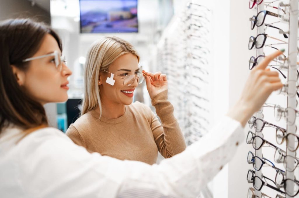 A young patient tries on a sample set of eyeglasses in store with their optometrist as a guide for a new prescription