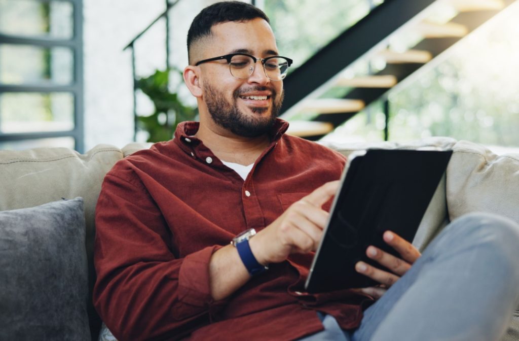 An image of a person sitting on the couch in their living room while wearing glasses and entertaining themselves on a tablet, they are happy and in a good mood.