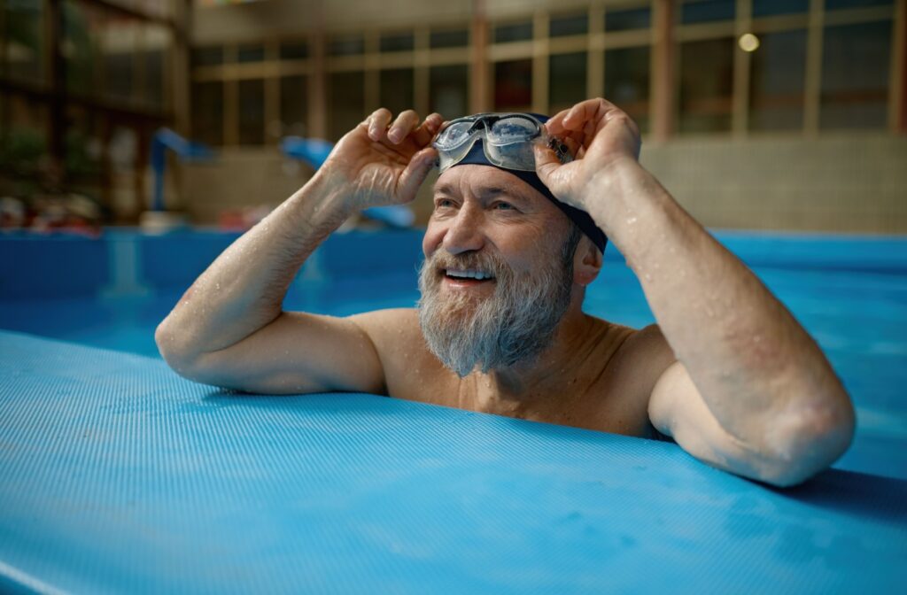 A person at the edge of a pool lifting up their prescription swim goggles while smiling.