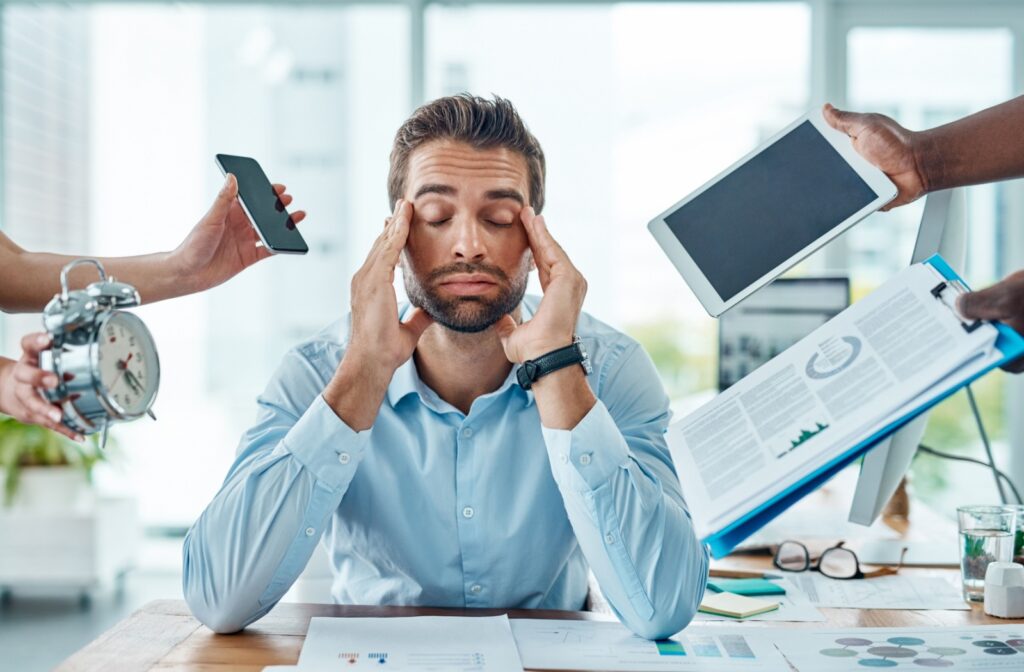 A man sitting at a desk with his eyes closed and hands on his temples, with hands holding out a clip board, phone, and an alarm clock, displaying the stress he is facing and how it's affecting his eyesight.