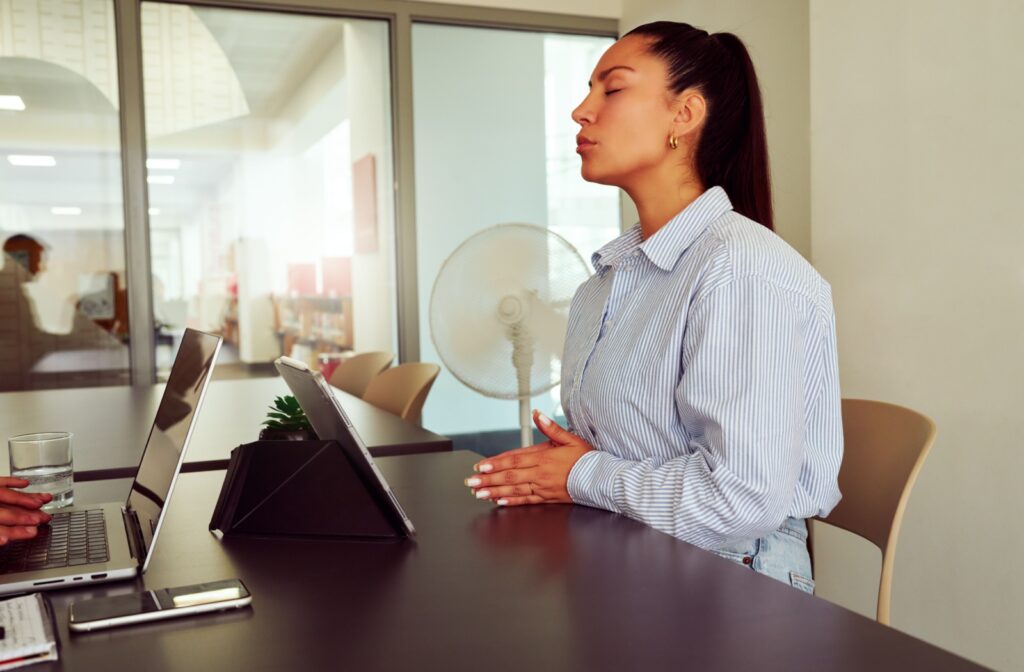 A woman sitting at a desk, eye closed, taking a deep breath to help reduce her stress that is leading to vision problems.